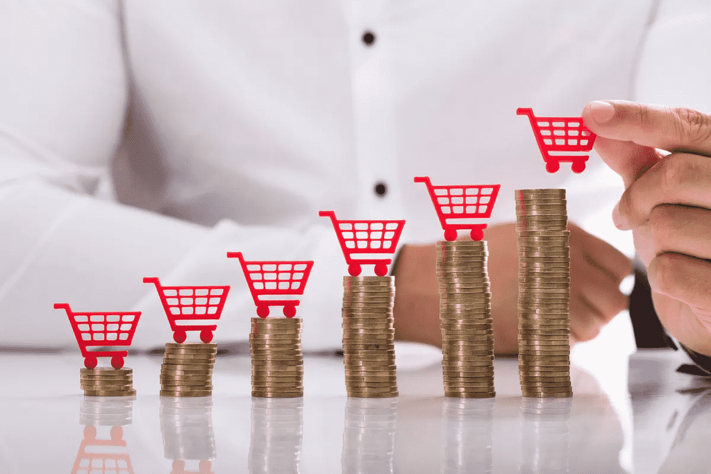 Mão colocando miniaturas de carrinhos de compras sobre pilhas de moedas cada vez mais altas, representando o aumento dos preços